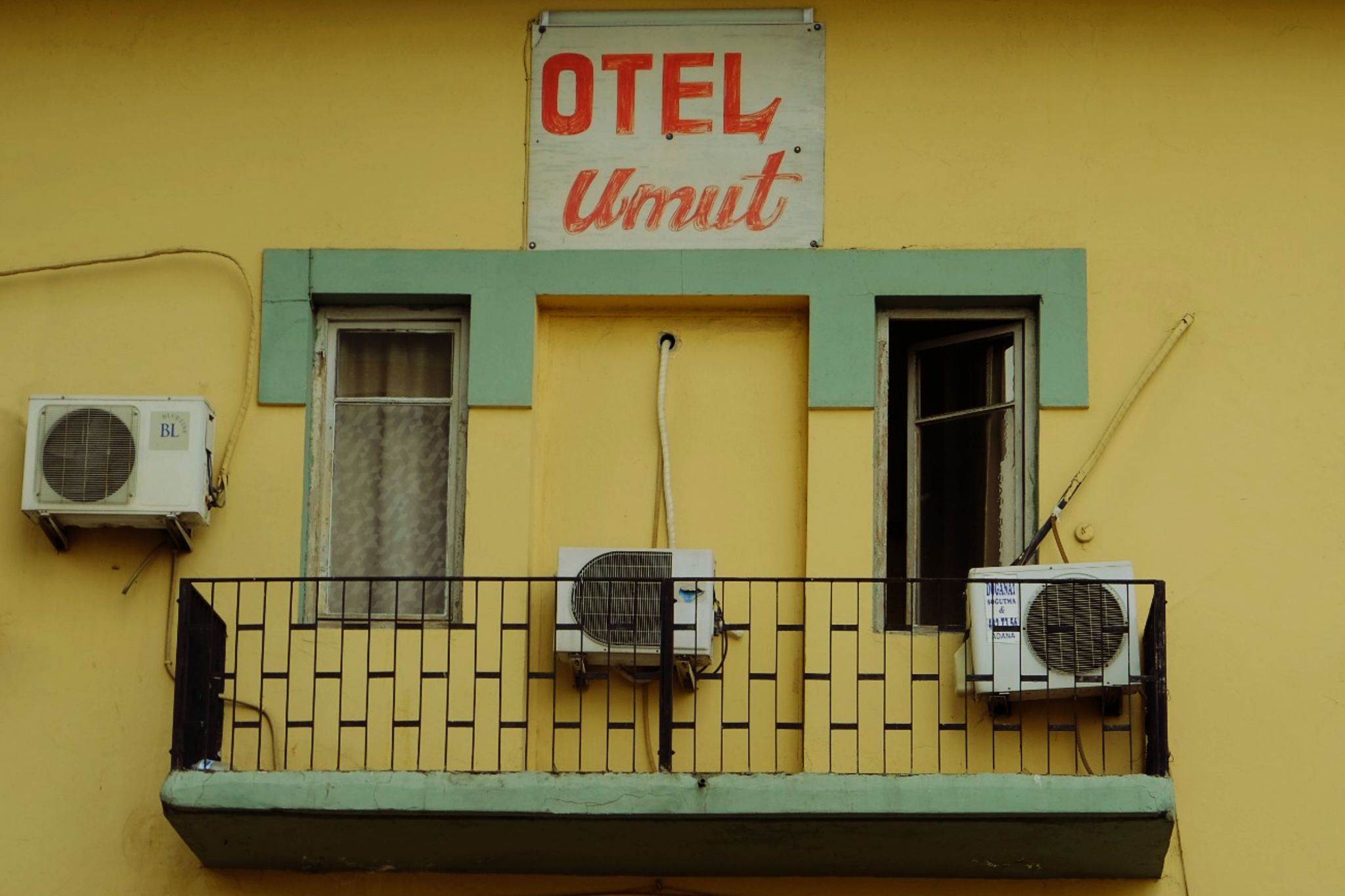 Aidiyet Fotoğrafçı: Esin Ruken Usuk Betimleyen: Alper OKÇUOĞLU (Çukurova Üniversitesi öğrencisi) Yatay dikdörtgen, renkli fotoğrafta “Otel Umut” tabelasının altında,  sarı renkteki otelin iki penceresinin açıldığı demir parmaklıklı balkonu yer alıyor.   Fotoğraf gündüz çekilmişçesine aydınlık. Balkonun altındaki boşluk ve pencerelerin demirsiz olması, aşağıda yer alan fotoğrafçının üst katta bir yeri fotoğraf kadrajına aldığını gösteriyor.  Fotoğrafın alt kısmının çoğunluğunu kaplayan bir balkon var. Balkonun zemini gri bir betondan oluşuyor. Balkonun siyah demir korkulukları, yaklaşık dikey olarak 10 adım uzunluğunda, yatay boşlukları yaklaşık 1 karış ve aralıklarında demirden yatay çubuklarla birleştirilmiş. Balkon 3 yönden demir korkulukla kapalı.  Fotoğrafa göre sokağa bakan taraf ile sağ ve sol tarafları kapalı. Fotoğrafta 3 adet klima dış ünitesi var.  Duvar üzerine sabitlenmiş iki klima dış ünitesi balkonun içinden duvara montajı yapılmış. Balkonun ortasında iki kolon dışarı çıkık şekilde duruyor. Balkon içinde olan dış ünitelerden biri kolonların ortasında, balkon zemininin yarım metre kadar üzerinde duruyor. Dış ünitenin klima ile hava bağlantısını ve elektrik akımını sağlayan kablolar dış ünitenin yaklaşık bir metre üzerinde duvara açılmış deliğin içinden geçirilmiş. İkinci dış ünite fotoğrafa göre balkonun sağ tarafına takılmış. İkinci ünitenin arkasında yani dış ünitenin 2 parmak kalınlığı kadar üstünde çevresi beyaz renkli tahtadan yapılmış cam yarı açık şekilde duruyor. İkinci dış ünitenin havalandırma ve elektrik kabloları sağ tarafa doğru yaklaşık 10 karış yukarı doğru giderek duvarın içinden geçirilmiş. Üçüncü dış ünite ekranın solunda balkonun dışında kalıyor. Bu dış ünitenin kabloları duvara yapışık şekilde kıvrılarak ünitenin üzerinden kadrajın dışına doğru gidiyor. Kolonların arasında yer alan klima dış ünitesinin sol tarafında pencere var. Pencere kapalı ve perdesi örtük. Perdenin rengi de tıpkı pencere gibi beyaz. Birinci ve ikinci dış üniteye yakın her iki pencerenin tahtaları soyulmuş bazı yerler sarı tahta izlerini ortaya çıkarıyor. Pencerelerin üstünden başlayarak her iki pencerenin sonuna kadar devan edecek şekilde yeşil renkte duvar boyanmış. Yeşil renk dört yerden aşağı doğru inecek şekilde boyanmış. İki uç kolonlardan diğer iki uçta pencere köşelerinden inecek şekilde boyanmış. Duvarın yeşil kısmının yani fotoğrafın yukarısında beyaz bir poster kağıdına kırmızı renkle OTEL Umut yazılı. Otel kelimesi büyük harflerle, daha belirgin ve kalın. Umut kelimesi daha silik şekilde ve sadece ilk harfi büyük. Umut kelimesinde harflerin bazı kısımlarında özellikle uçlarında kıvrımlı ve bitişik şekilde yazılmış.