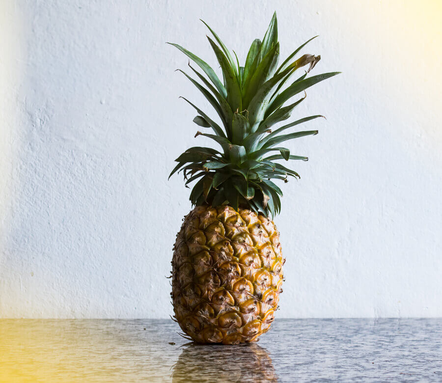 Ananas Betimleyen: Nilay VARDAR  Fotoğrafta yer alan, gri tezgah üzerine dikey şekilde konmuş ananas meyvesi, iki karış yüksekliğe ve yarım karış ene sahip. Meyve, sert görünümlü bir gövde ile onun üzerine konumlanmış yeşil yapraklardan oluşuyor. Gövdenin, alta ve üste doğru ovalleşen dikey dikdörtgenimsi bir yapısı var. Kahverengi-sarı tonlardaki dış kabuk, dikey elips şeklinde küçük parçalara ayrılmış. Bu parçaların orta kısmından, dışarı doğru sarkan 1 santimetreden kısa otsu yapılar görülüyor. Meyvenin yapraklı bölümünün alt kısımlarında aşağı sarkan, orta bölümden itibaren yukarı doğru yükselen ucu sivri yeşil yapraklar var.