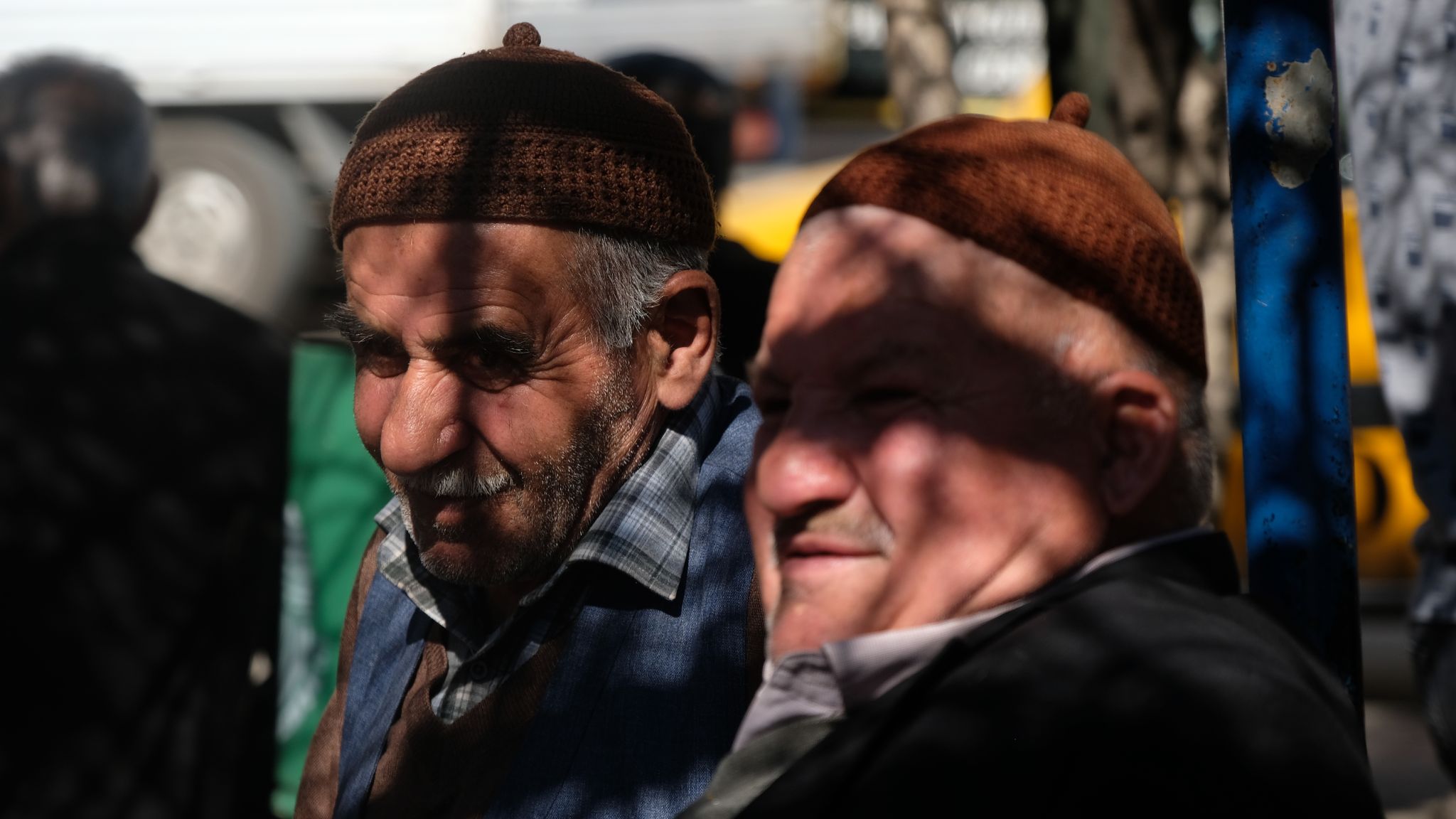 Bakış Renkli enine dikdörtgen fotoğrafta , 50 ve 60’lı yaşların arasında iki tane erkek, açık alanda, güneşli bir günde yan yana oturuyor. Hafif sağlarına dönükler ve fotoğrafın soluna bakıyorlar. Arka plan bulanık.  Kameranın önündeki iki erkekten birisi kameraya daha yakın diğeri uzak kalıyor. Yakın olan erkeğin sadece omzundan yukarısı kadraja alınmış, yüzünü sola dönmüş, sırtını mavi ince bir direğe yaslamış. Açık kahverengi, ucunda fındık kadar ponponu olan şapka giymiş. Yüzü tombul ve elmacık kemiği belirgin. Gözlerinin üstüne gölge düşmüş, hafif kahverengimsi kaşlarını çatmış, gözlerini kısarak uzağa bakıyor. Göz kenarlarının etrafında kırışıklıkları belirginleşmiş. Burnu büyük ve burun ucuna doğru genişliyor. Dudağı küçük ve ince. Uçlarına doğru beyazlaşmış ince bir bıyığı var. Ensesindeki görünen şaçları kısa ve beyaz. Grimsi gömleğinin yakası boynunu kapatmış, gömleğinin üzerine siyah ceket giymiş.  Objektife uzak kalan, yanındaki erkeğin ise göğsünden yukarısı kadraja alınmış, yüzünü sola dönmüş. Koyu kahverengi, ucunda fındık kadar ponponu olan şapka giymiş. Yüzü ince, uzun ve elmacık kemiği belirgin. Siyah, içinde hafif beyazları olan dağınık kaşları var. Gözlerinin altı torbalı ve uzağa bakıyor. Burnu uzun, tombul ve geniş, burnunun ucundan dudağının kenarına doğru bir çizgisi var. Alnında ve göz çevresindeki kırışıklıkları belirgin. Sakalı ve bıyığı beyaz. Alt dudağı ince, üst dudağını bıyığı kapatmış, görünmüyor. Saçlarının sadece kulak üstündeki küçük bir kısmı görünüyor ve beyaz renkte. Üzerinde kareli, siyahımsı çizgileri olan gömlek, gömleğin üstünde kahverengi renkte kazak ve kolsuz mavimsi bir ceket giymiş. Fotoğrafın bulanık olan arka kısmında iki tane daha erkek var. İkisi de arkasını dönmüş ayakta ve birinin sadece kafası birinin ise belden yukarısı ve beyaz saçları görünüyor.