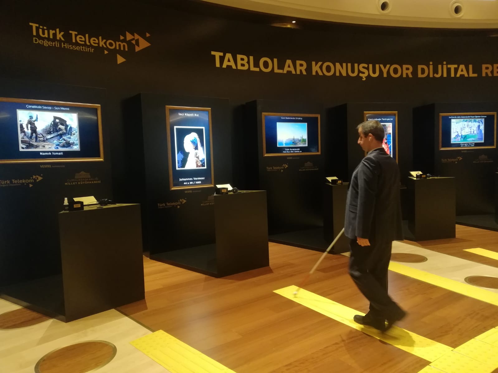 Yatay dikdörtgen renkli fotoğrafta “Tablolar Konuşuyor dijital re” yazısının ve Türk Telekom değerli hissettirir yazılı logonun yer aldığı siyah duvardaki ekranlarda dört tablo ve bu tablolara doğru duran Kenan Önalan yer alıyor. Kapalı mekanın yerleri açık ahşap parkeli. Sarı bir ışık ortama loş hava vermiş. Fotoğrafın solundan sağına doğru sıralanan tabloların önünde bel hizasına dek uzanan masalar ve üzerinde küçük tabela ve kulaklıklar duruyor. Tablolar fotoğrafta çok net değil. Kenan Önalan koyu renk takım elbiseli 40’larında, kısa siyah saçlı. Sağ elinde beyaz baston ile yere döşenmiş sarı renkli kılavuz iz de tablolara doğru adım atar halde. Sol profilinden fotoğrafta yer alıyor, hareket halinde olduğu için flu şekilde.