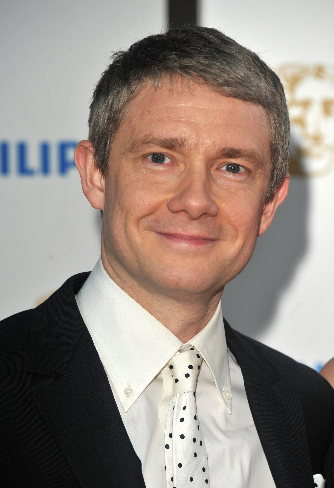 Oyuncu Martin Freeman Betimleyen: Gökçe ALTINÇEKİÇ (Boğaziçi Üniversitesi, ED200 dersi öğrencisi)  Dikey dikdörtgen ve renkli olan fotoğraf karesinde Martin Freeman’ın sadece göğsünden üst kısmı kadraja alınmış. Freeman’ın bedeni hafif sola doğru dönse de yüzü direkt kameraya bakıyor. Freeman, fotoğrafta kırklı yaşlarında görünüyor.  Kare bir yüz yapısına sahip olan Freeman’ın hafif çekik ve küçük sayılmayan grimsi renkte gözleri var ve göz altı torbaları belirgin. Kahverengi, kalın ve dağınık olan kaşları gözlerine yakın. İnce ve biraz yamuk bir burun duvarına sahip olan Freeman’ın burun kanatları ve delikleri geniş ve sivri olmayan dik bir burun ucuna sahip. Geniş olan alnının ortasında tüm alnına yatay şekilde uzanan iki tane alın çizgisi bulunuyor. Gülümsediği için elmacık kemikleri daha da belirginleşmiş ve dudaklarının ve burnunun çevresinde çizgiler oluşmakla birlikte iki yanağında da gamzeler ortaya çıkmış. Pembe, düz ve ince olan dudaklarından üstteki alttakinden biraz daha ince. Freeman; geniş ve sivri olmayan bir çeneye, büyük, hafif kepçe kulaklara ve yapışık kulak memesine sahip. Ayrıca sakalı ya da bıyığı bulunmuyor. Kısa, sık ve düz saçlarını sol tarafına doğru yatıran Freeman, saç diplerinden uçlarına doğru siyahtan griye bir renk farkı görülüyor. Ayrıca Freeman’ın alnının sağ kısmında, burun direğinde, sağ yanağında ve çenesinin orta kısmının sağ tarafında küçük boyutlarda açık kahverengi benler bulunuyor. Kısa ve kalın olan boynunda adem elması belirgin. Aynı zamanda Freeman’ın, yüzünden normal bir kiloya sahip olduğu anlaşılıyor.  Boynunun alt kısmını kaplayan, sivri uçlarında birer küçük ve beyaz düğmeleri olan geniş yakalı beyaz, düz bir gömlek giyen Freeman, küçük bir düğümle bağlanmış, siyah küçük benekleri olan beyaz bir kravat takmış. Gömleğinin yakası yüzünden boynunun sağında bir çizgi oluşmuş. Gömleğinin üstüne siyah bir ceket giyiyor ve ceketin sol göğüs tarafında kadraja tamamen sığmayan kravatıyla aynı desende olan bir mendil bulunuyor. Sağ omzunun büyük bir kısmı fotoğraf karesinde görünmüyor fakat sol taraftaki geniş omzu görünüyor.  Arka planda beyaz duvar üzerinde Freeman’ın sağ tarafında ve kulaklarının hizasında flu, mavi renkte yazılar yazıyor. Sol tarafında kalan kısımda ise Freeman’ın saçlarının hizasında kalan tarafta flu, altın rengi bir insan kafası sembolü ve aşağıya doğru giden Freeman’ın kafasının gölgesi görülüyor.