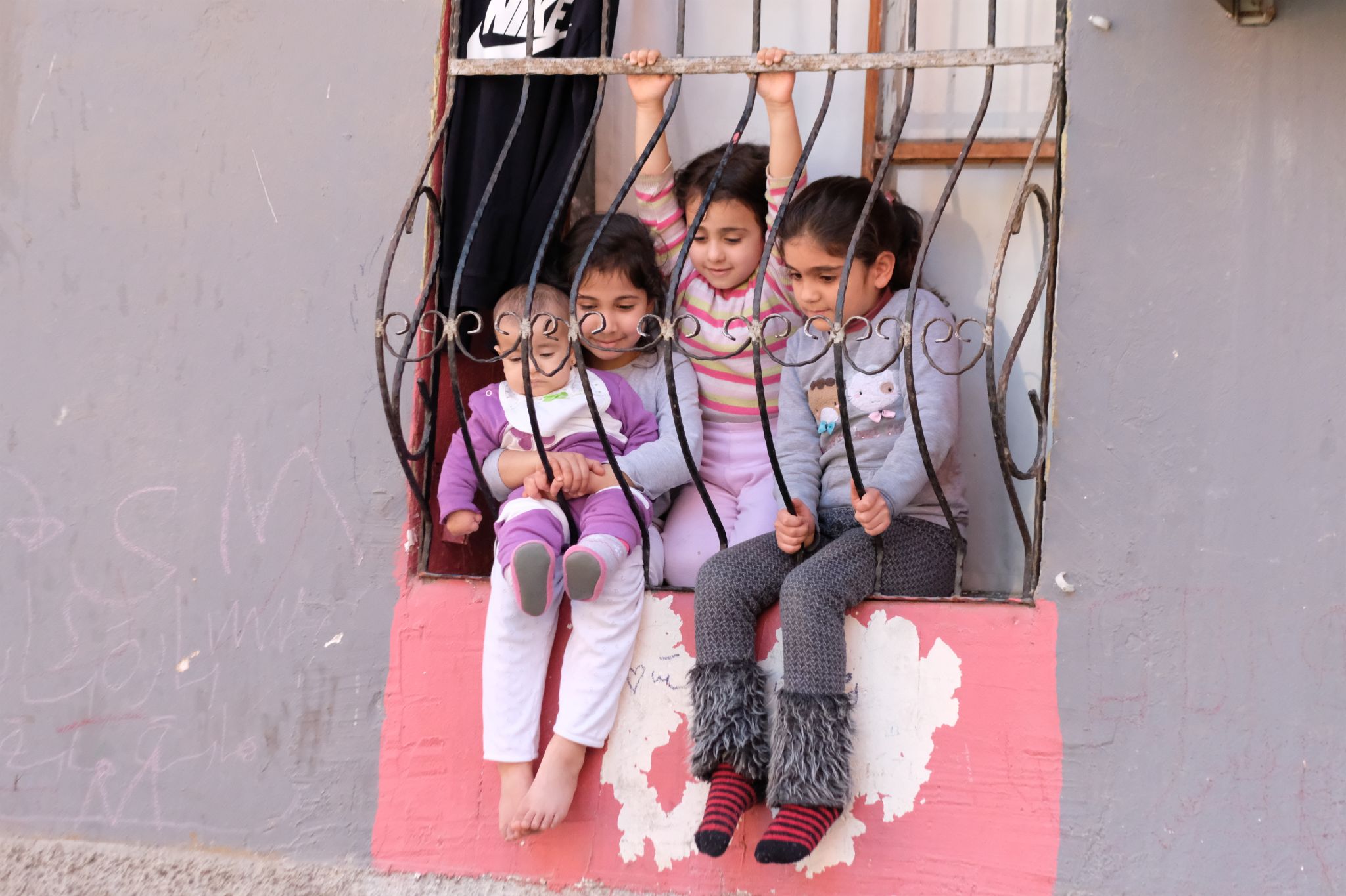 Safderun Yatay dikdörtgen renkli fotoğrafta gündüz vakti bir evin parmaklı penceresinde oturup ayaklarını sarkıtan üç çocuk ve bir bebek yer alıyor. Pencerenin solunda 7-8 yaşlarında bir kız çocuğu ve kucağında birkaç aylık bir bebek var. Kız çocuğunun saçları arkadan toplu ve pencereden aşağı bir noktaya bakıyor. Saçları siyah, alnı dar, kirpikleri siyah kaşları düz, burnu küçük, ten rengi beyaz, dudakları ince, üzerinde mavi bir kazak ve beyaz bir eşofman altı var. Ayakları çıplak,pencereden sarkıyor. Sol ayağı sağ ayağının üstünü kapatıyor.  İki eliyle bebeğin belinden tutarak bebeği sarıyor. Bebek beyaz tenli çok az saçı var yüzünün bir kısmını pencere demirleri kapatıyor.  Bebek de yere kızın baktığı noktaya bakıyor. Bebeğin üzerinde beyaz bir mama önlüğü var üzerinde yeşil motifler var , mor bir kıyafet giymiş. Bebek kıyafetinin diz kısmında beyaz çizgiler var. Ayağında pembe bir ev patiği ve pembe patiğinin alt gri kısımları görünüyor. Bebek fotoğrafa göre sol elini yumruk yapıyor. Ayakları pencereden sarkıyor.  Kucağında bebek olan kızın hemen yanında bitişik şekilde oturan 7-8 yaşlarında başka bir kız çocuğu var. Siyah saçlı dar alınlı beyaz tenli . O da bebekle ve diğer kızla birlikte aynı yere bakıyor. Gri,pembe, yeşil çizgiler olan  bir kazak giymiş. Elleriyle üstten demirleri tutuyor ve dizlerinin üstünde oturuyor. Altında pembe bir eşofman var.  Bu kızın hemen yanında 9-10 yaşlarında başka bir kız çocuğu duruyor. Bu kız çocuğunun saçları siyah arkadan at kuyruğu yapılmış şekilde toplu, diğer çocuklarla aynı noktaya bakıyor. Gözleri siyah, teni buğday rengi. Üstünde üzerinde tavşan motifleri olan gri bir kazak giymiş. Elleri ile demirin altını tutuyor. Açık gri üzerine koyu gri motifleri olan bir pijaması  ve ayak bileğinin dizinin yarısına kadar tüylü bileğe giyilen çorapları var. Kırmızı siyah çizgili çorap giymiş. Ayakları demirden aşağı sarkıyor. Diğer çocuklarla aynı yere bakıyor. Fotoğraftaki duvar lila renginde  ve üzerine tebeşirle yazılmış yazılar var.  Fotoğrafın solunda pencerenin üst kısmında siyah bir kumaş parçası sarkıyor ve kumaşın üzerinde nike yazıyor. Pencerenin arkasında beyaz bir fon perdesi var. Demirlerin arkasında kahverengi, ahşap, T şeklinde pencere çerçevesi var.  Duvarın ortasında yarım görünen demirli bir pencere mevcut. Demirli pencerenin altında lila duvarın ortasında kalan soyulmuş pembe bir boya yer alıyor. Pencerenin demirleri siyah. Lila, pembe duvar  bitiminin  alt kısmında gri beton  pürüzlü bir duvar var.