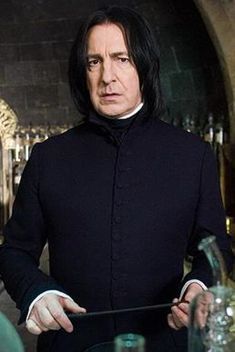 Harry Potter And The Philosopher`s Stone / Harry Potter Felsefe Taşı Filmi -Severus Snape- Betimleyen: Sedef TAYLAN /  Boğaziçi Üniversitesi ED 200 Dersi Öğrencisi  Dikey dikdörtgen fotoğrafın ortasında belden yukarısı gözüken Severus Snape (Alan Rickman) iki eliyle asasının uçlarından tutarak ayakta duruyor. Arka planda büyük gri tuğlalardan oluşan kubbe şeklinde bir tavan gözüküyor. Altın sarısı detayları olan duvarda cam şişeler bulunuyor.  Severus Snape ellili yaşlarının ortasında görünen beyaz tenli bir erkek. Siyah, parlak ve düz saçları omuz hizasının biraz yukarısında son buluyor. Kulakları saçlarının arkasında kalmış. Geniş bir alnı ve çenesi kemikli oval bir yüzü var. Siyah kaşları hafif çatılmış ve iki kaşının ortasında bir çizgi belirmiş. Küçük kahverengi gözlerinin altında göz altı torbaları belirmiş. Gözleri endişeli bir şekilde yere doğru bakıyor. Uzun ve kemikli burnunun kenarlarından dudak çevresine doğru derin çizgiler gözüküyor. Üst dudağı alt dudağından daha ince. Soluk renkte dudakları hafif aralık ve düz bir biçimde duruyor. Kafasını hafif sağa doğru çevirerek poz vermiş. Çenesinde ve dudak kenarlarında kırışıklıklar var.  Boğazlı, siyah renkte ve dar bir kaban giymiş. Kabanın altında, boynundan ve kol manşetlerinden görünen beyaz renkli gömlek tarzı bir kıyafet var. Sağ eliyle siyah renkli, düz asasını kavramış, diğer eliyle ise asanının diğeri ucunu tutmuş. Snape’in önünde bulanık bir biçimde cam şişelerin tepe kısımları gözüküyor.
