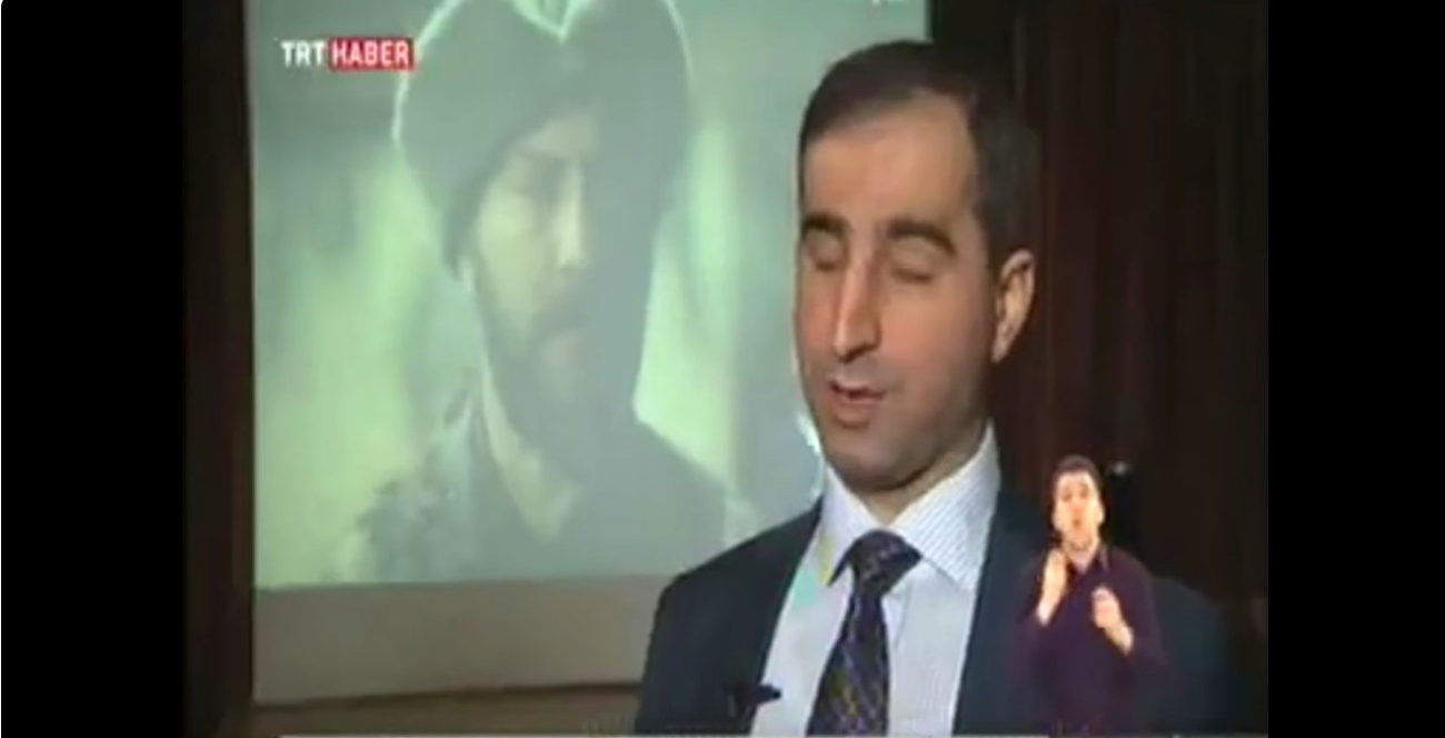 Kenan Önalan TRT Haber ekranında konuşurken sağ alttaki işaret dili çevirmeni konuşulanları çeviriyor. Önalan'ın gerisinde duran ekranda bir tarihî dizi karakteri yer alıyor.