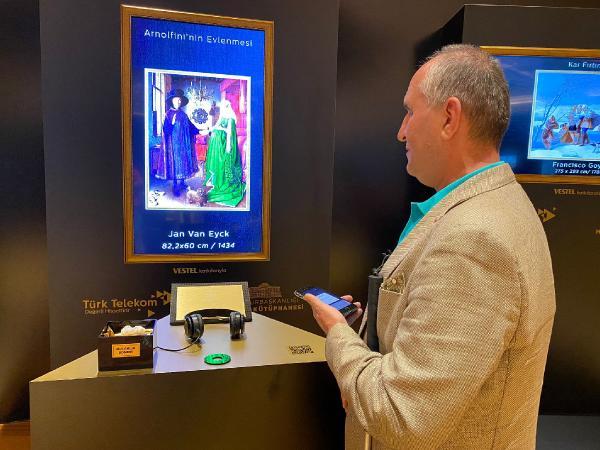 Krem rengi ceketli Olgun Yılmaz Dijital Resim Sergisi'nde, elindeki telefonla Jan Van Eyck'in Arnolfi'nin Evlenmesi adlı tablosunun önünde duruyor.
