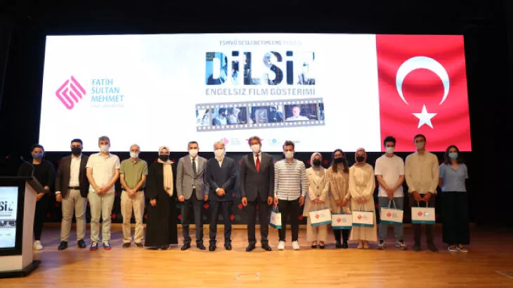 Öğrenciler engelsiz bir yaşam için el ele verdiler Yatay dikdörtgen fotoğrafta etkinlik katılımcılarının sahnede yan yana poz verdiği görülüyor. 15 katılımcıdan altısı kadın, dokuzu erkek. Poz verilen sahnenin arkasındaki büyük ekranın solunda Türk bayrağı, ortada büyük harflerle FSMVÜ SESLİ BETİMLEME yazısı yer alıyor. Bu yazının hemen altında çok daha büyük punto ile DİLSİZ yazısı mevcut. Ekranın en solunda ise logosuyla birlikte Fatih Sultan Mehmet Vakıf Üniversitesi yazısı yer almakta.
