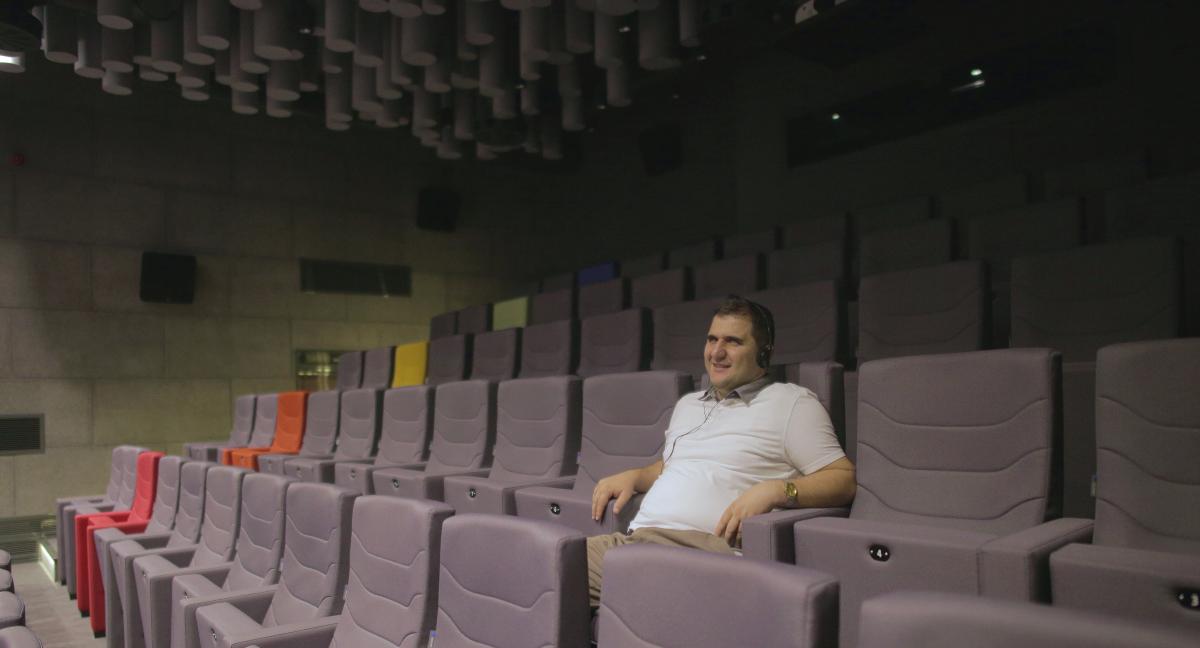 SineBU’da Sesli Betimleme SineBU sinema salonunda çekilen fotoğrafta Engin Yılmaz koltuklardan birinde oturmuş tebessüm ediyor. Kulağında taç kulaklığı var.